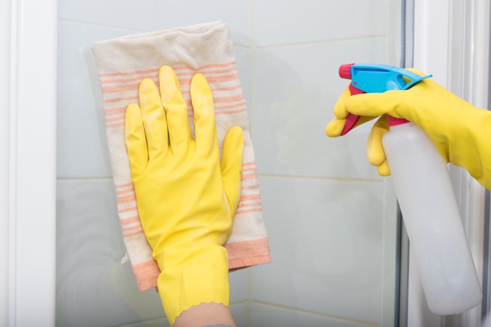 ゴム手袋をした手で浴室のガラス扉を布で拭き、スプレーを持つ様子