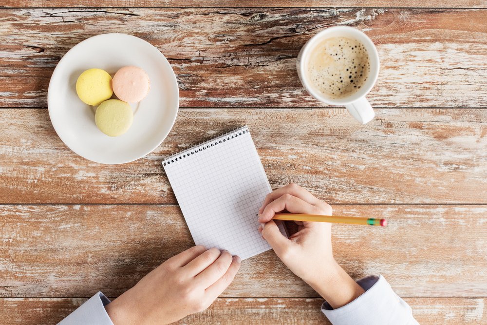 木のテーブルでメモ帳に書き込む手元とコーヒー、焼き菓子